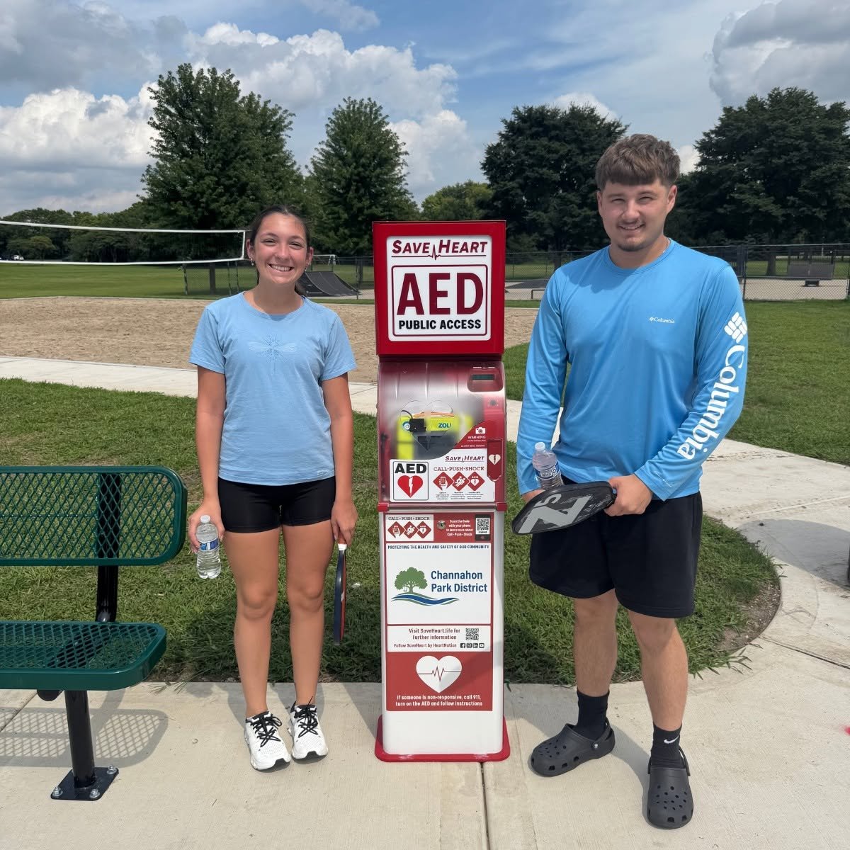 Two pickleball players beside a new SaveHeart by HeartNation Tower with the Channahon Park District sponsor panel at Central Park in Channahon, Illinois